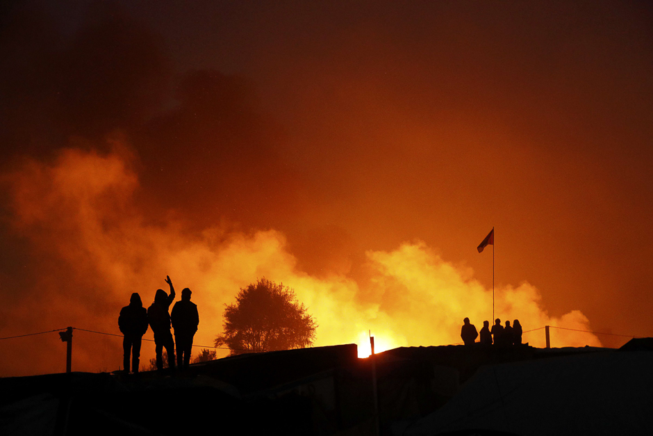 Migrants are seen in silhouette as they gather near flames from a burning makeshift shelter on the second day of the evacuation of migrants and their transfer to reception centers in France, as part of the dismantlement of the camp called the 
