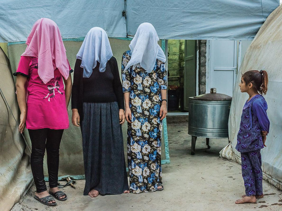 A girl watches as three Kurdish women are photographed with their faces hidden. Two of the women say they were forced to marry ISIS fighters before escaping to a refugee camp. PHOTO: YURI KOZYREV/NATIONAL GEOGRAPHIC