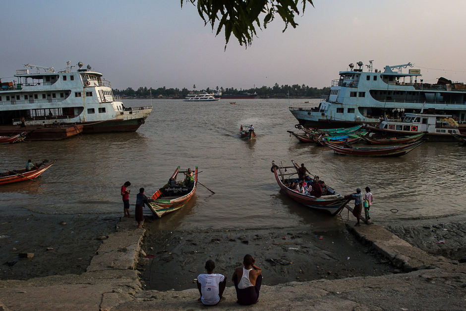 This picture taken shows men watching as people commute by boat from a jetty in Yangon. PHOTO: AFP