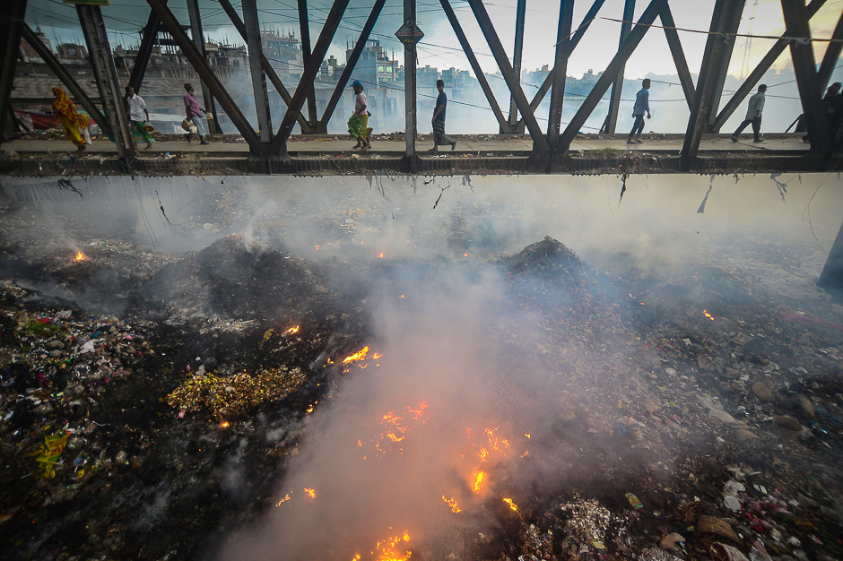 Bangladeshi pedestrians cross an iron bridge as smoke rises from a fire in a garbage dump below them near the Buriganga river in Dhaka. PHOTO: AFP