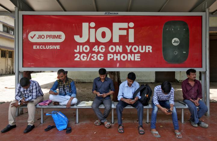 commuters use their mobile phones as they wait at a bus stop with an advertisement of reliance industries 039 jio telecoms unit in mumbai india july 10 2017 photo reuters