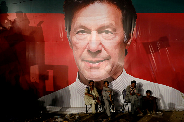 Labourers, who set up the venue, sit under a wall with a billboard displaying a photo of Imran Khan. PHOTO:REUTERS