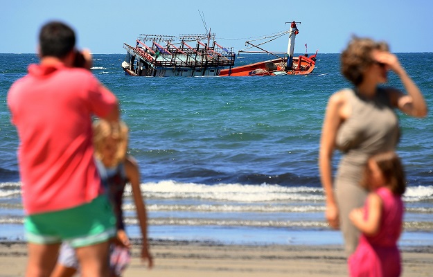Locals stand on a beach in front of a sunken vessel. PHOTO: REUTERS