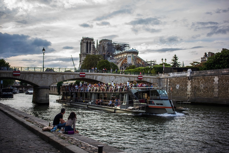 A couple of persons sit on the Seine river banks as a fly boat cruises by the Notre-Dame de Paris' Cathedral in Paris. PHOTO: AFP