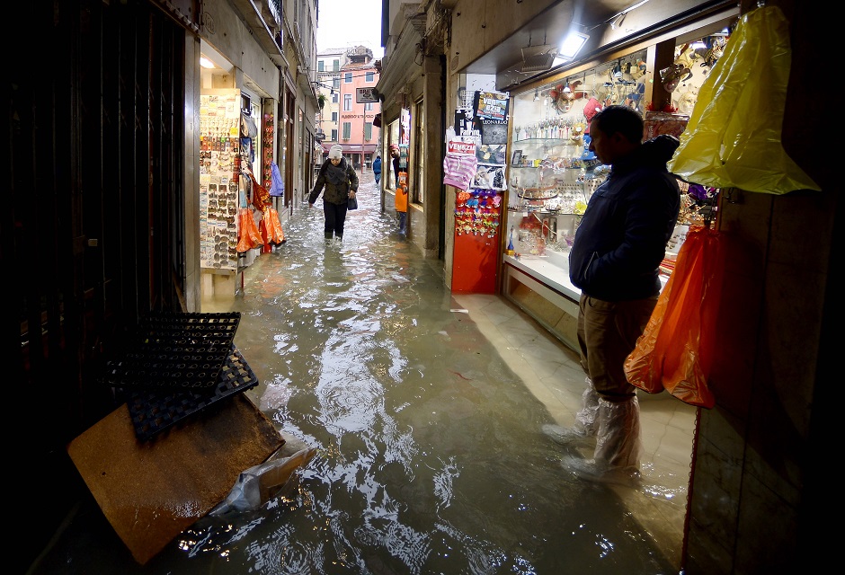 A man stands outside a souvenirs shop as a woman walks across a flooded alleyway. PHOTO: AFP